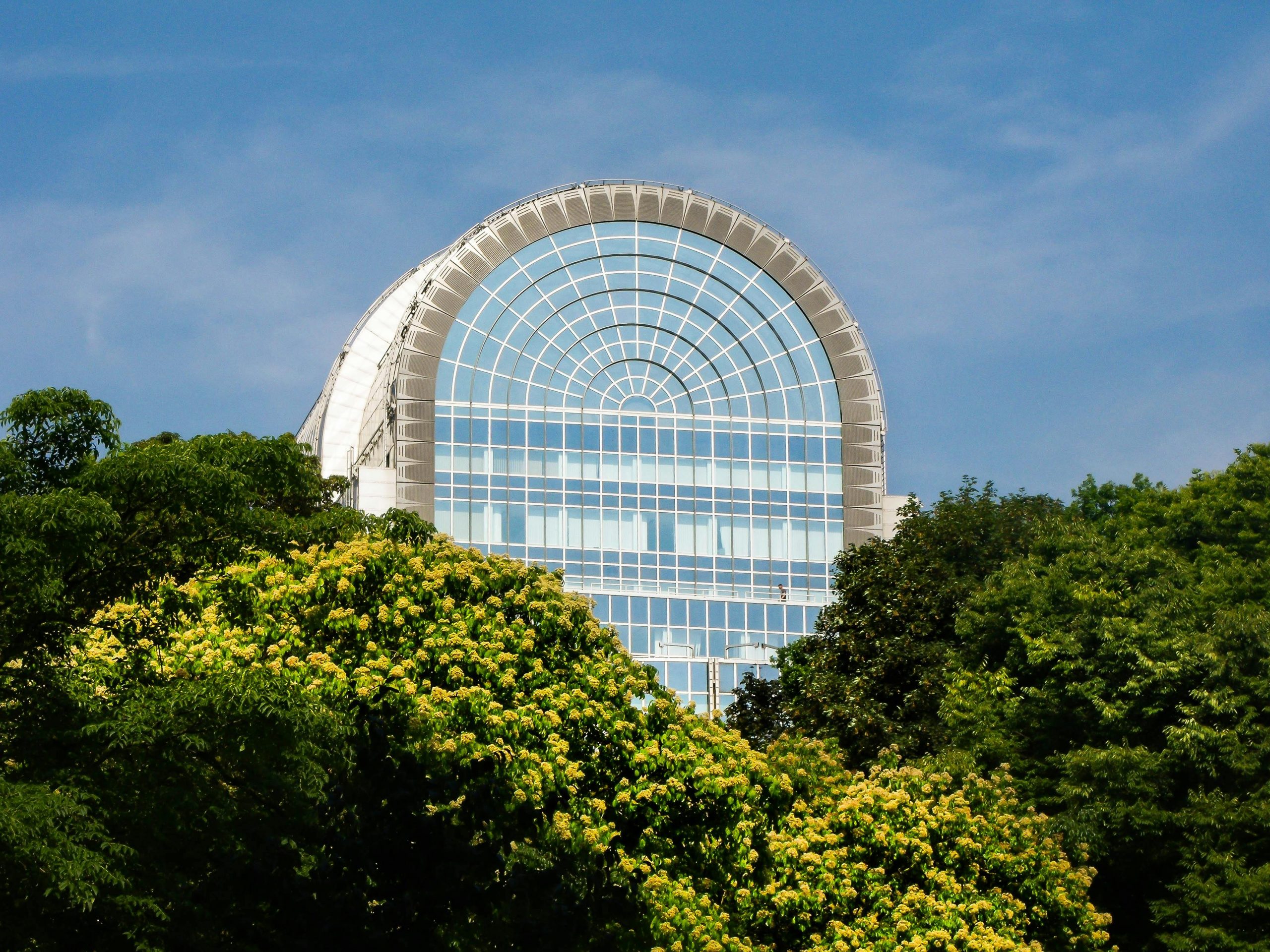 The curved glass rooftop of the Paul-Henri Spaak building of the European Parliament in Brussels, seen rising above the flowering trees of Parc Léopold on a clear summer day.